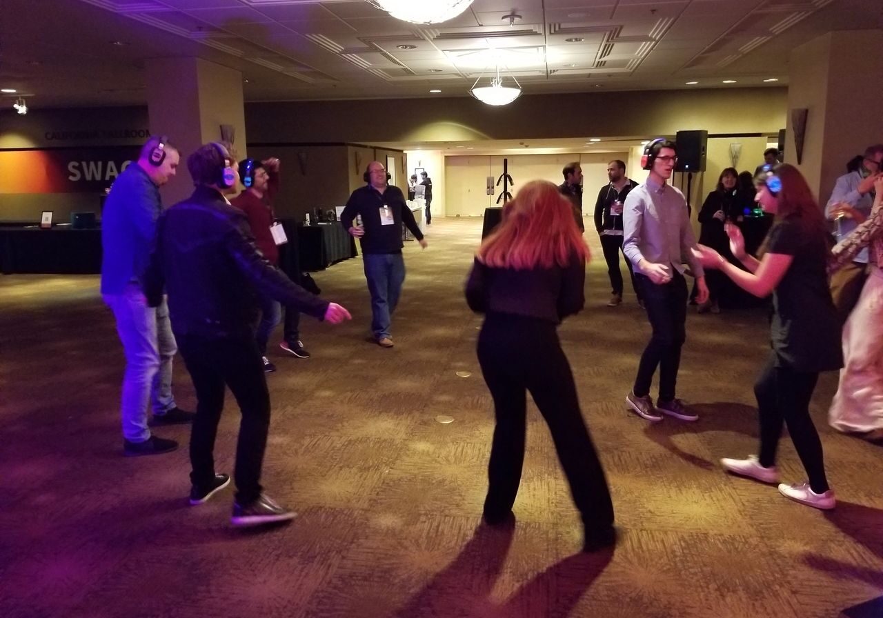People wearing headphones dance in a dimly lit room, participating in a silent disco event. Other attendees stand or watch in the background.