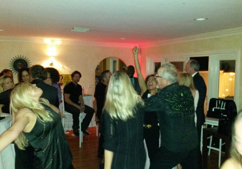 A group of people dance and socialize in a dimly lit room at a party; drinks are visible on a nearby table.