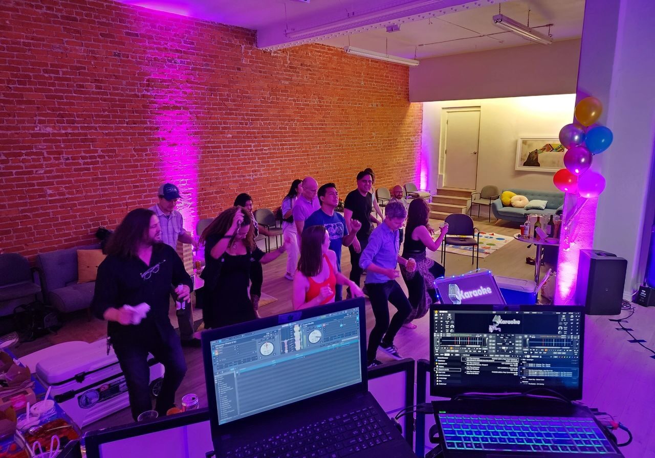 A group of people dance in a room with exposed brick walls and purple lighting, viewed from behind DJ equipment. Balloons and snacks are visible in the background.