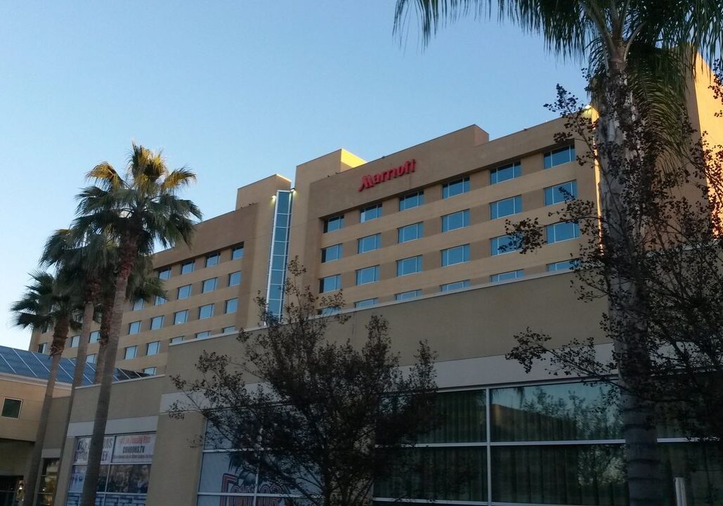 A beige multistory Marriott hotel building with palm trees in front, viewed from ground level under a clear blue sky.