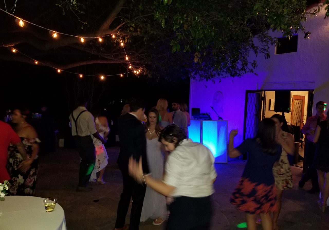 dj services, los angeles dj services People dance outdoors at night under string lights, with a lit DJ booth and a white building in the background. Drinks and tables are visible in the scene.