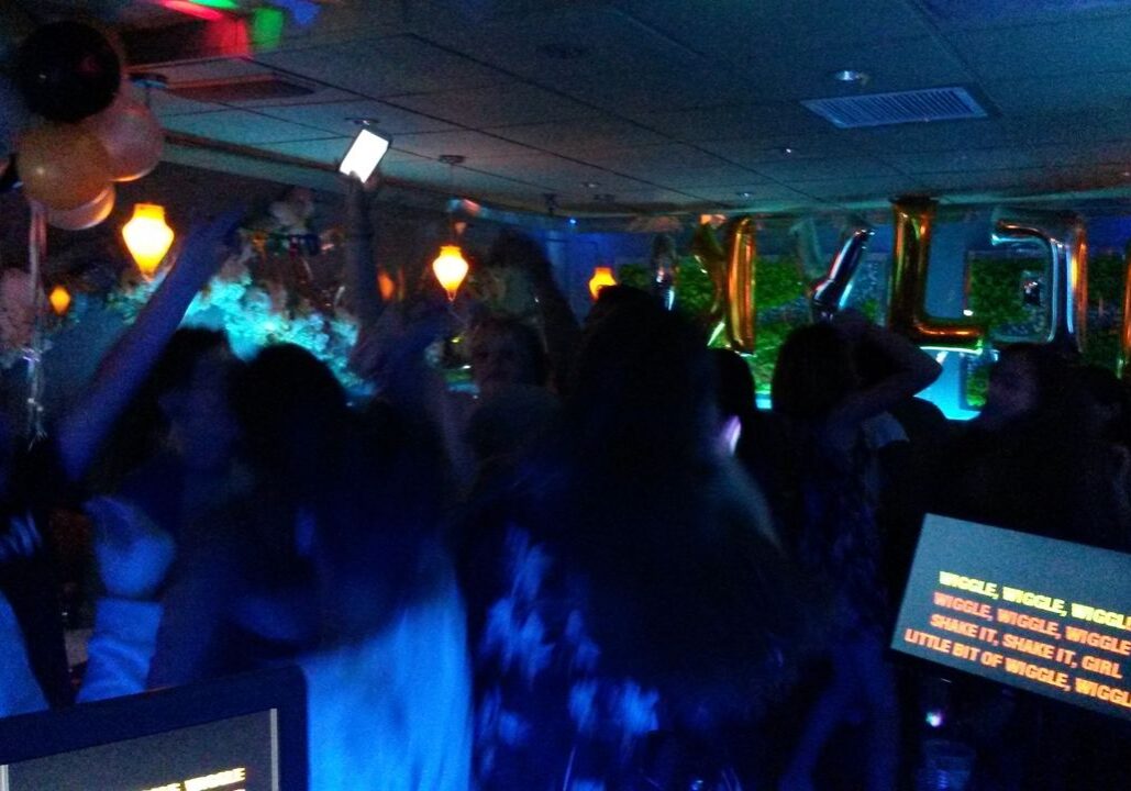 A group of people dance in a dimly lit room decorated with balloons and gold letter balloons, while karaoke lyrics are displayed on a screen in the foreground.