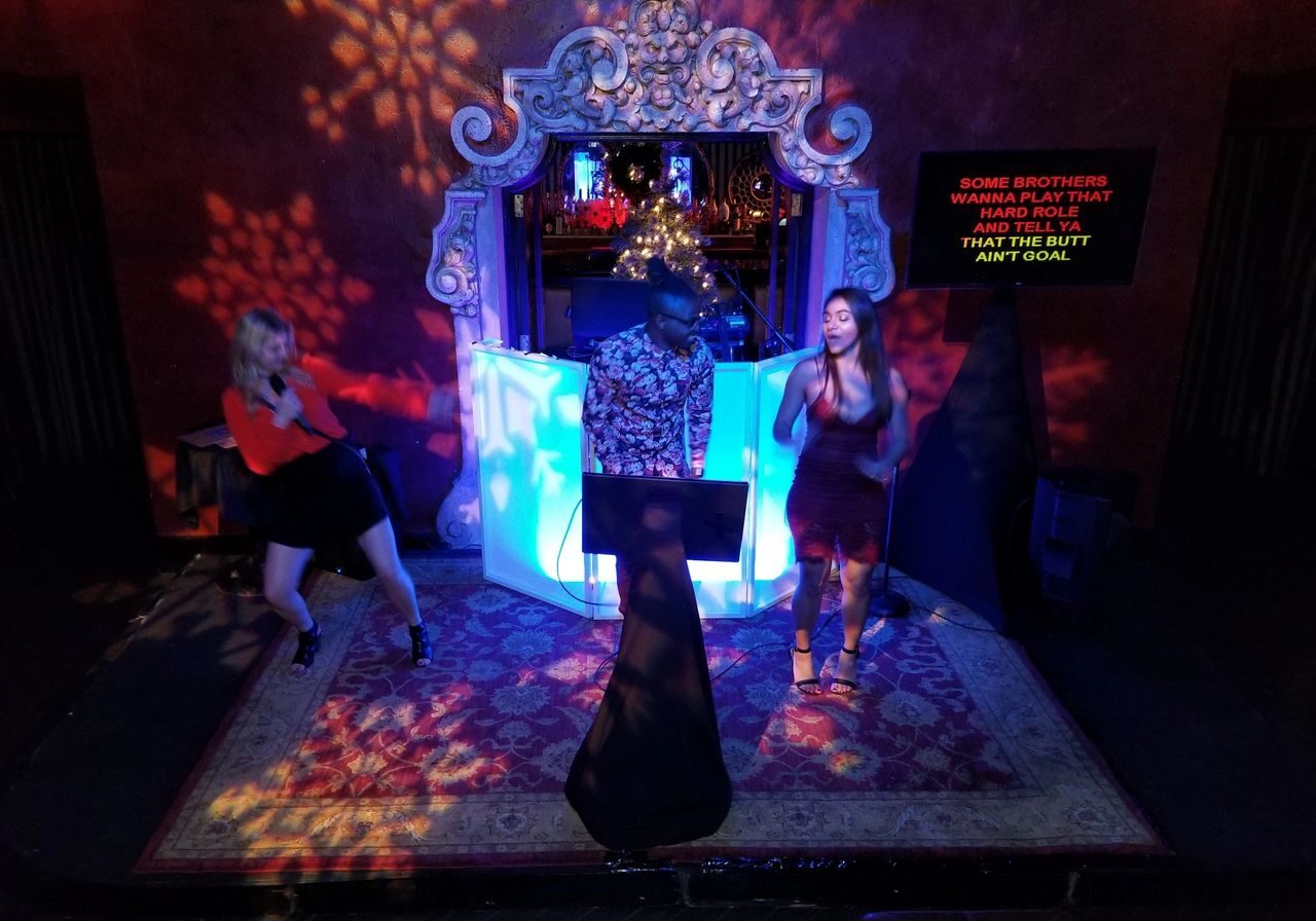 Three people are performing on a small stage with a decorative backdrop and colorful lights; lyrics are displayed on a screen to the right.