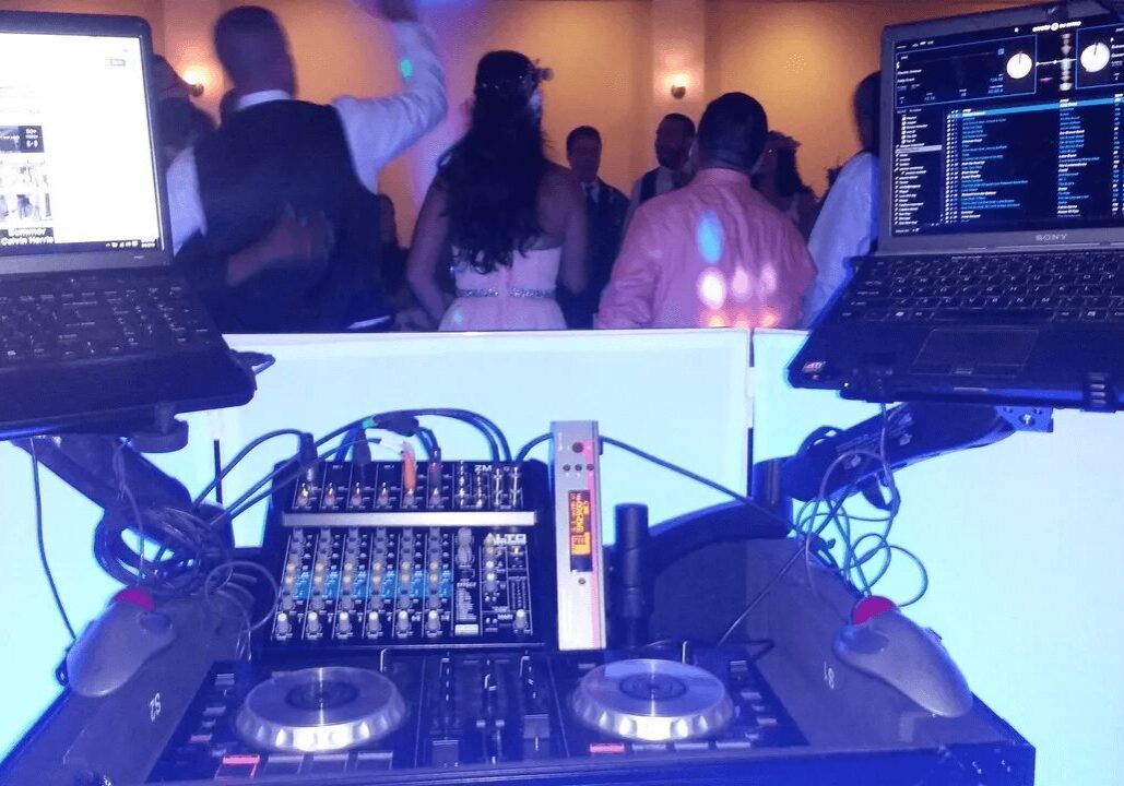 DJ setup with two laptops, a mixer, and turntables at a party with people dancing in the background.