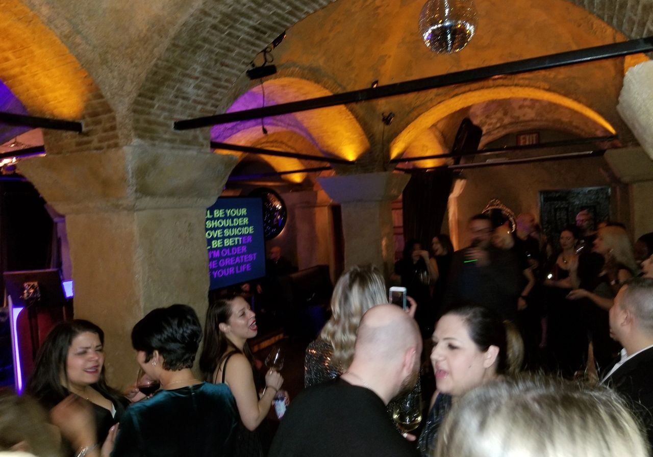 A group of people socializes at a dimly lit indoor party with arched ceilings and a disco ball; a karaoke screen displays song lyrics in the background.