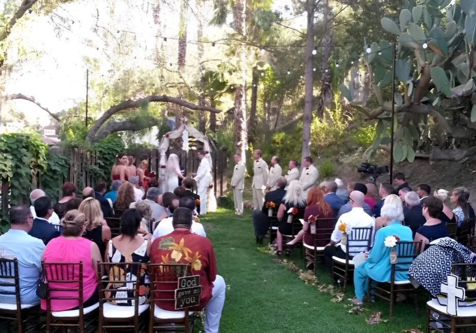 Outdoor wedding ceremony with a bride and groom under an arch, surrounded by a wedding party in matching attire. Guests are seated on either side in a garden setting with trees and lights.