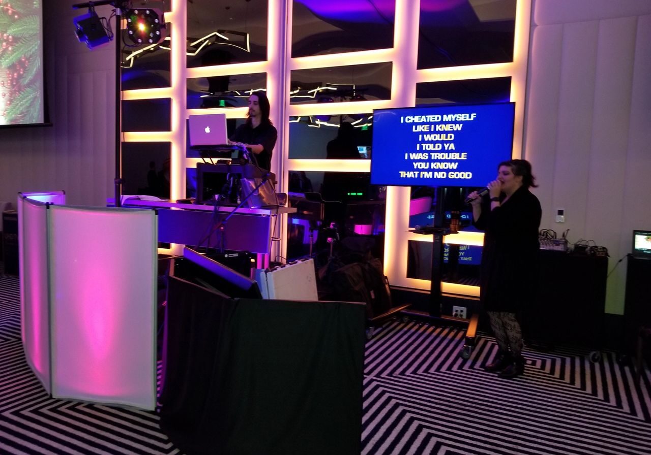 A woman sings into a microphone while another operates a laptop and DJ equipment; karaoke lyrics are displayed on a large screen behind them.