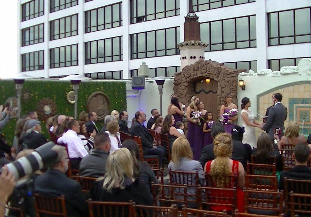 Outdoor wedding ceremony with guests seated, a wedding party in front, and an officiant marrying a couple near a decorative wall with a city building in the background.