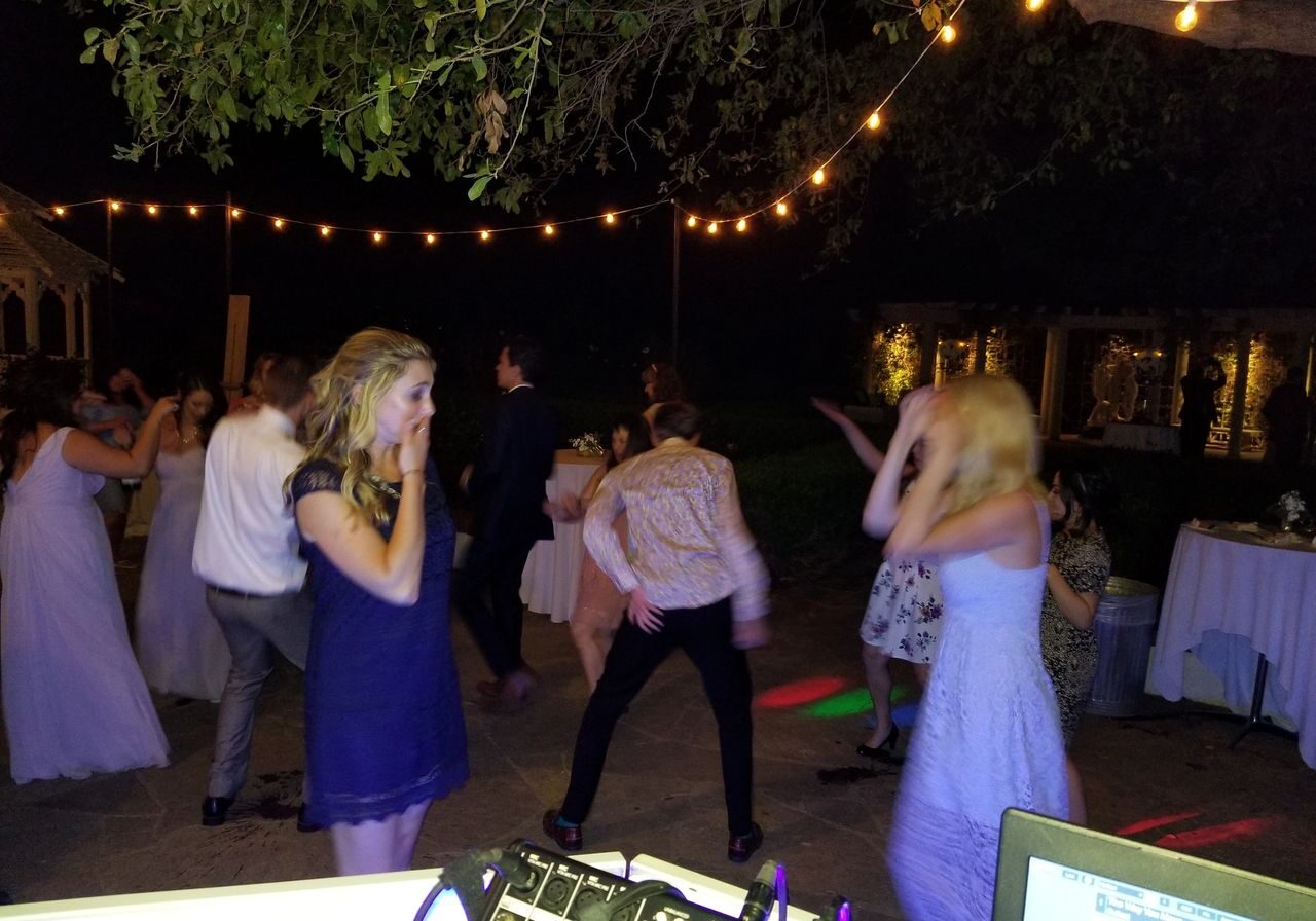 People dancing outdoors at night under string lights, with DJ equipment visible in the foreground and tables in the background.