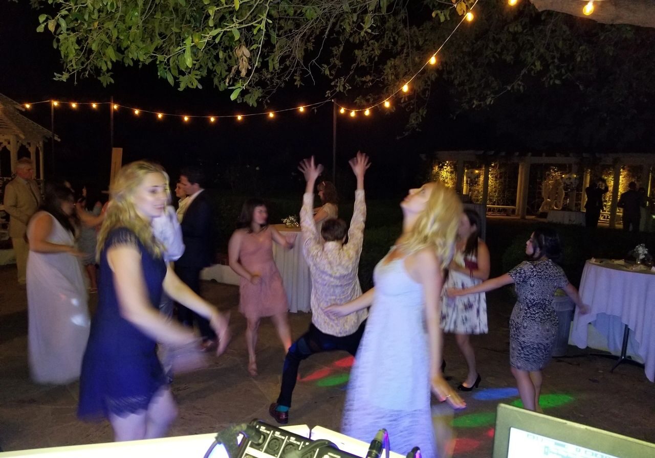 A group of people dance energetically on an outdoor patio at night, surrounded by string lights, with tables and trees in the background.