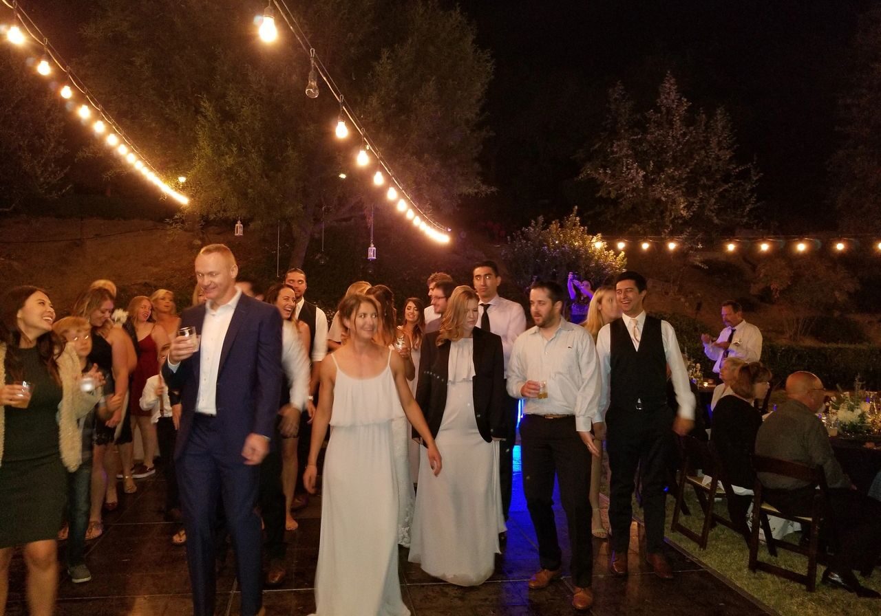 A group of people in formal attire walk together on a lit outdoor floor at night, with string lights overhead and trees in the background.