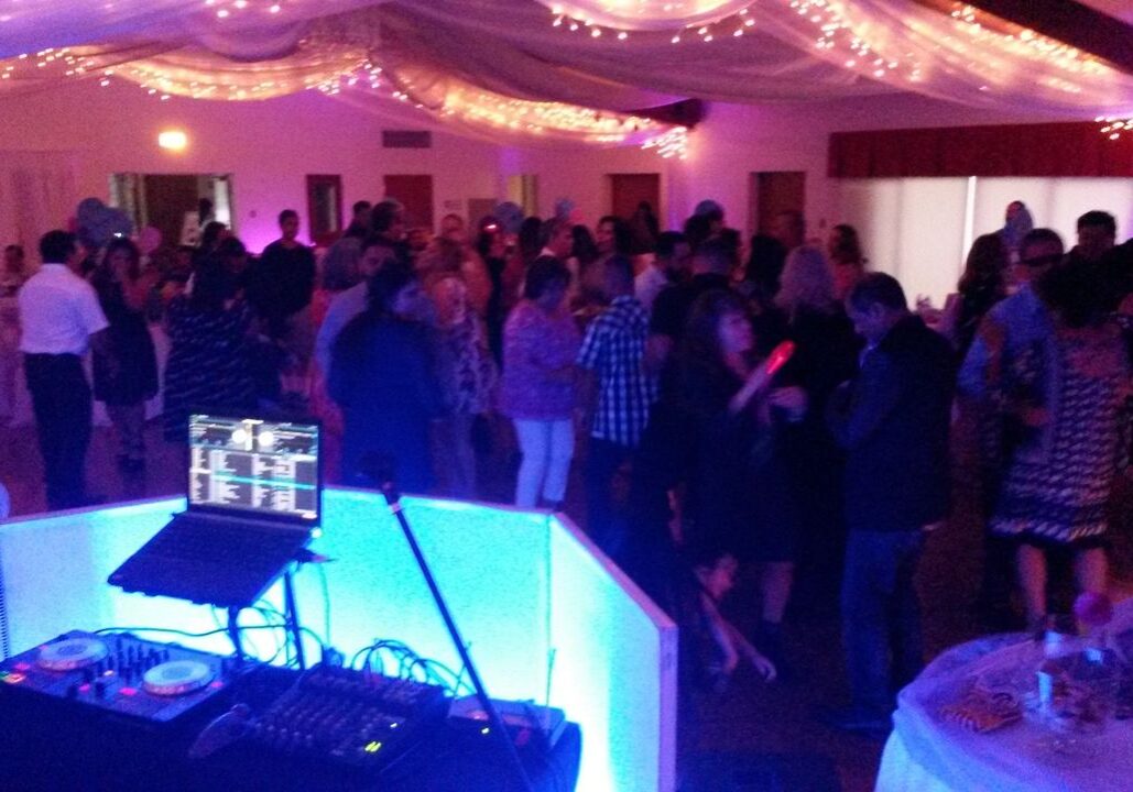 A large group of people are dancing in a decorated banquet hall with string lights, viewed from behind a DJ booth with turntables and a laptop.