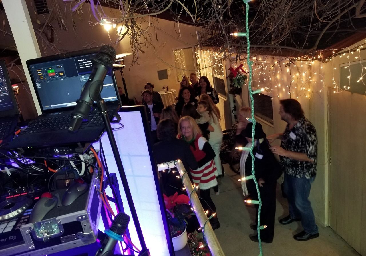 A group of people socialize and dance outdoors at night near a DJ booth, surrounded by string lights and festive decorations.