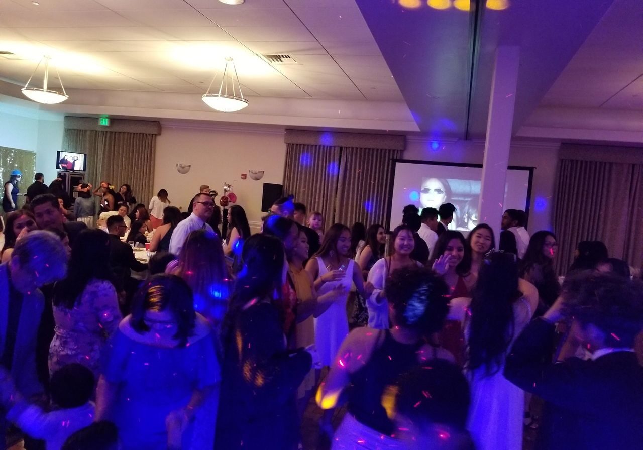 A large group of people are dancing and socializing in a banquet hall with colorful lights and a projection screen in the background.