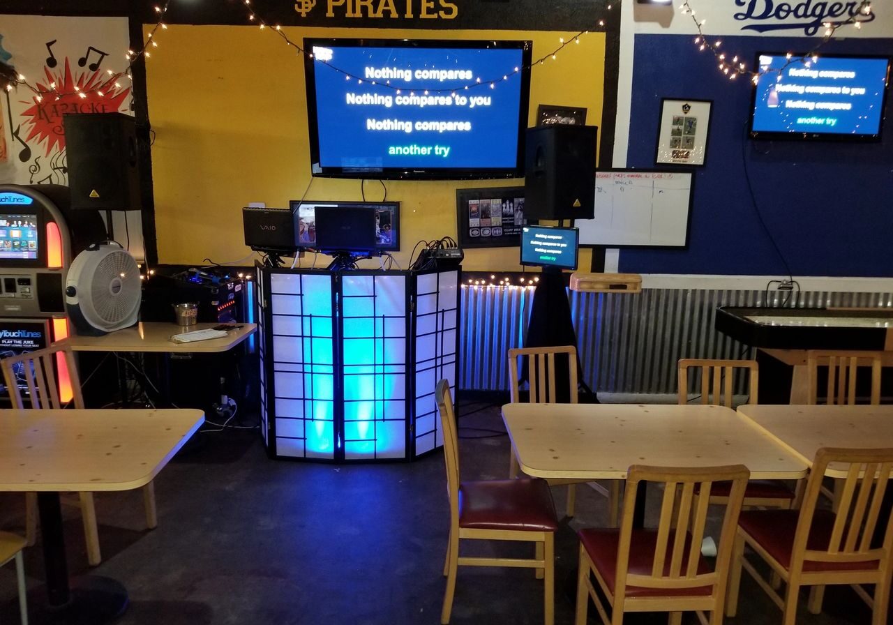 A karaoke setup in a casual dining area with lyrics on TV screens, wooden tables and chairs, and sports team signs on the walls.