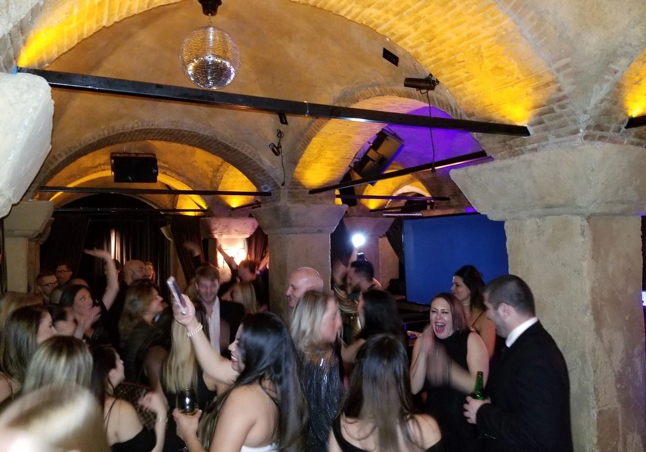 A group of people dressed in formal attire are dancing and socializing at a lively party in a stone-arched room with yellow lighting and a disco ball.