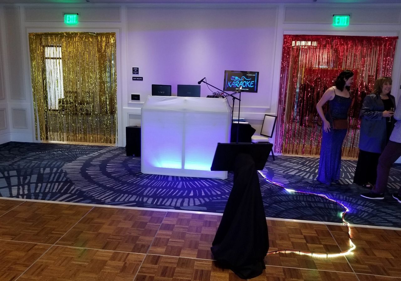 A karaoke setup with a lit DJ table, screen, and microphone stand on a dance floor; gold and red metallic streamers hang over two doorways; a few people stand nearby.