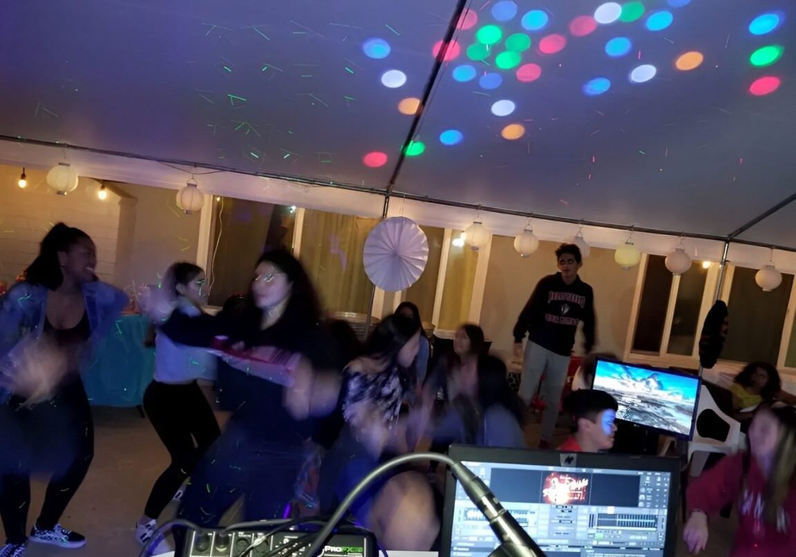 People are dancing under a tent with colorful lights and a DJ setup visible in the foreground.