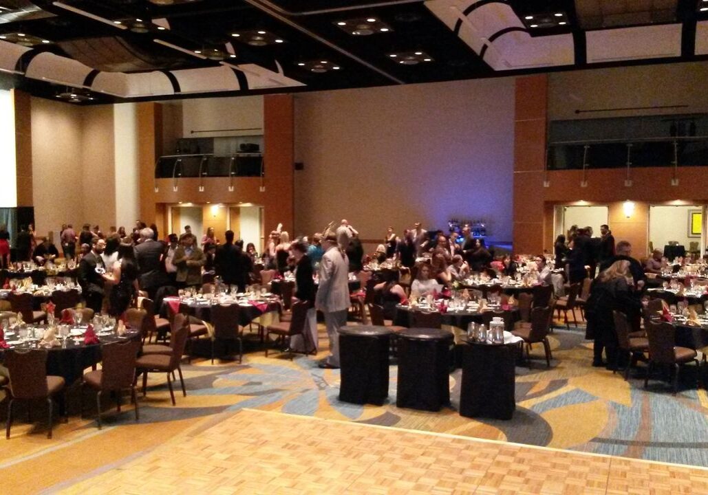 People gather and socialize around banquet tables in a large, carpeted event hall with a wooden dance floor in the foreground.