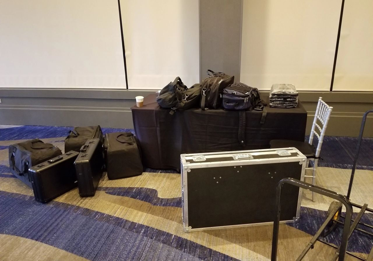 A table and floor hold several black equipment cases, bags, folded cloths, and a cup; a folding chair is nearby on a blue-patterned carpet.