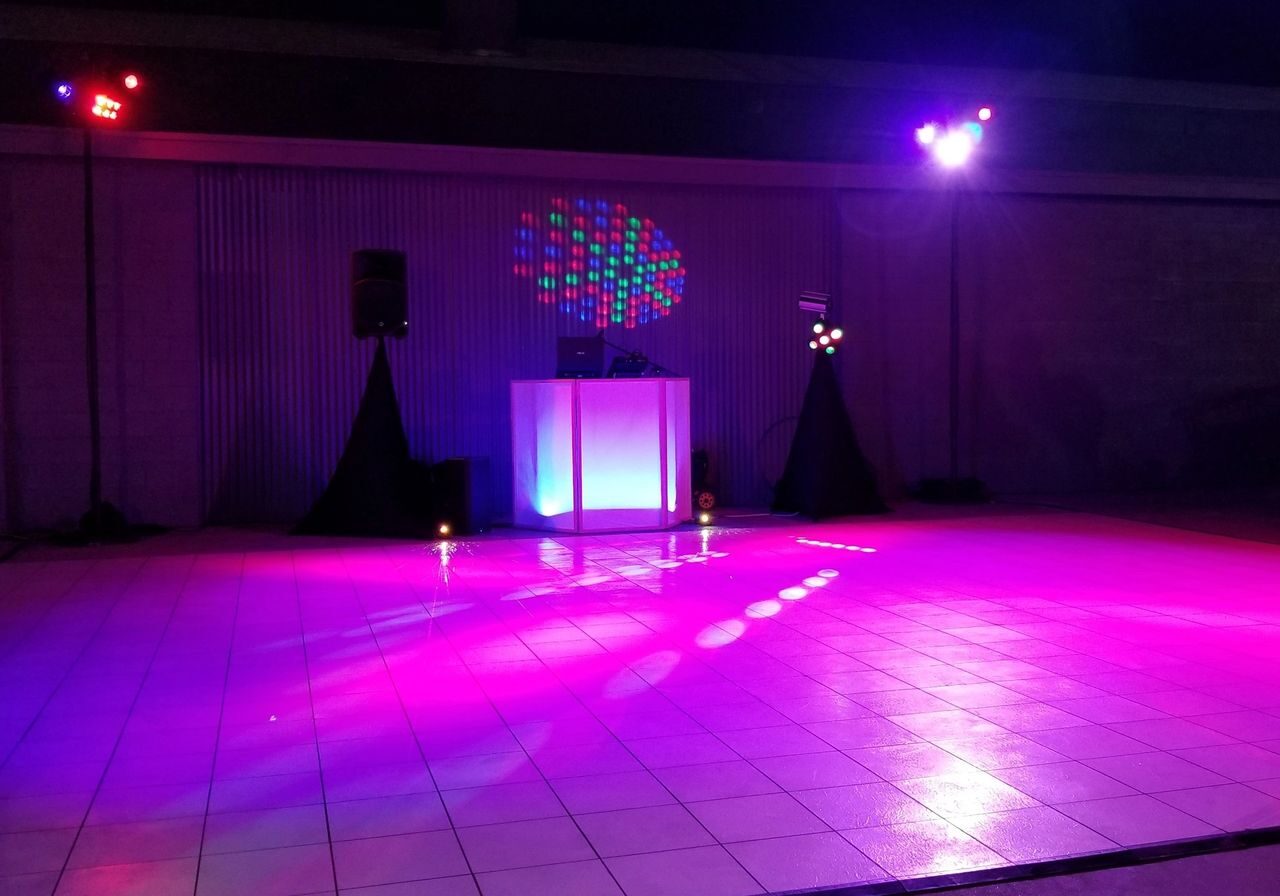 A DJ booth with speakers and colorful lights is set up on a tiled dance floor in a dimly lit indoor space.