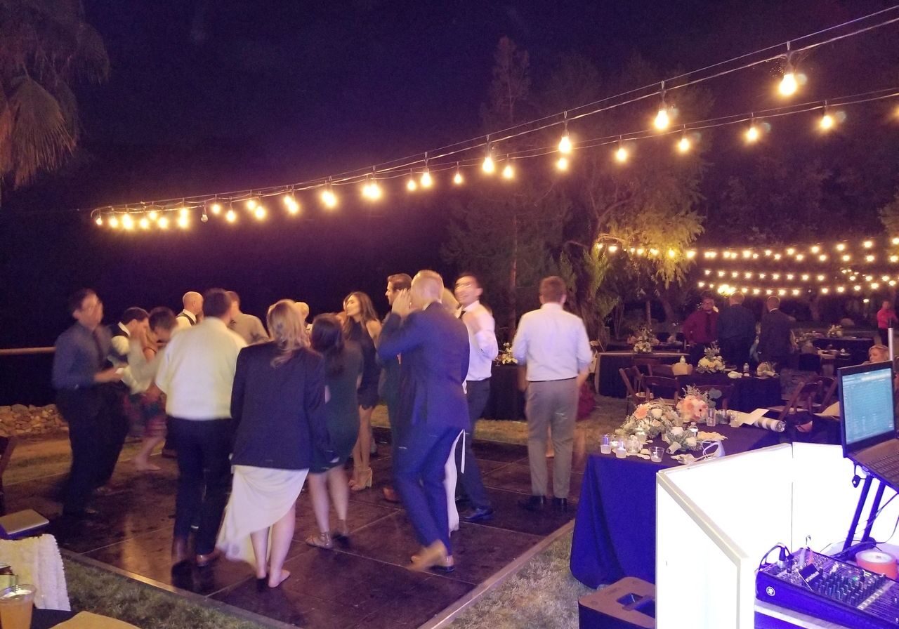 A group of people are dancing on an outdoor dance floor at night under string lights, with tables and a DJ booth nearby.