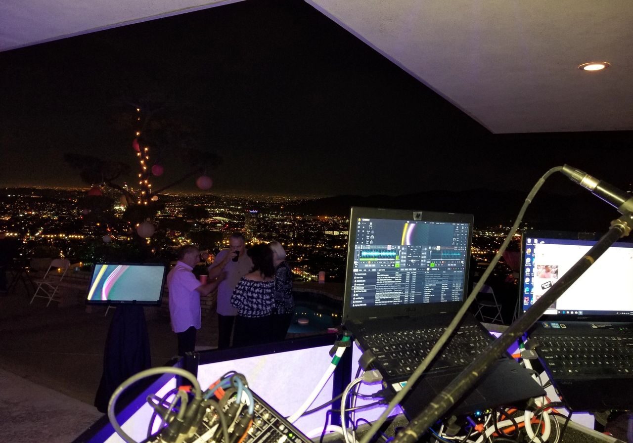 A nighttime rooftop scene shows DJ equipment in the foreground, with people dancing and city lights visible in the background.