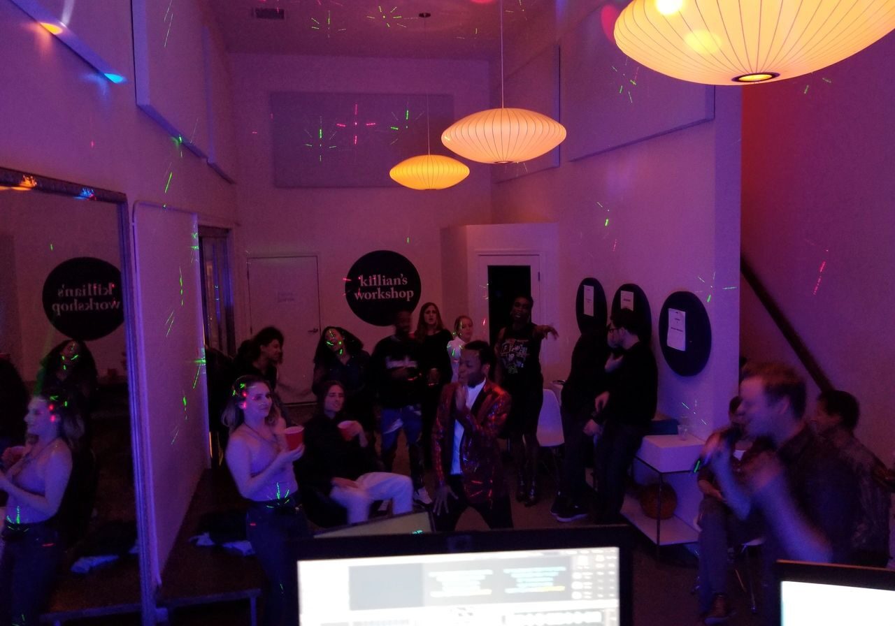 A group of people socialize and dance in a dimly lit room with colorful lights and hanging lamps; computer monitors are visible in the foreground.