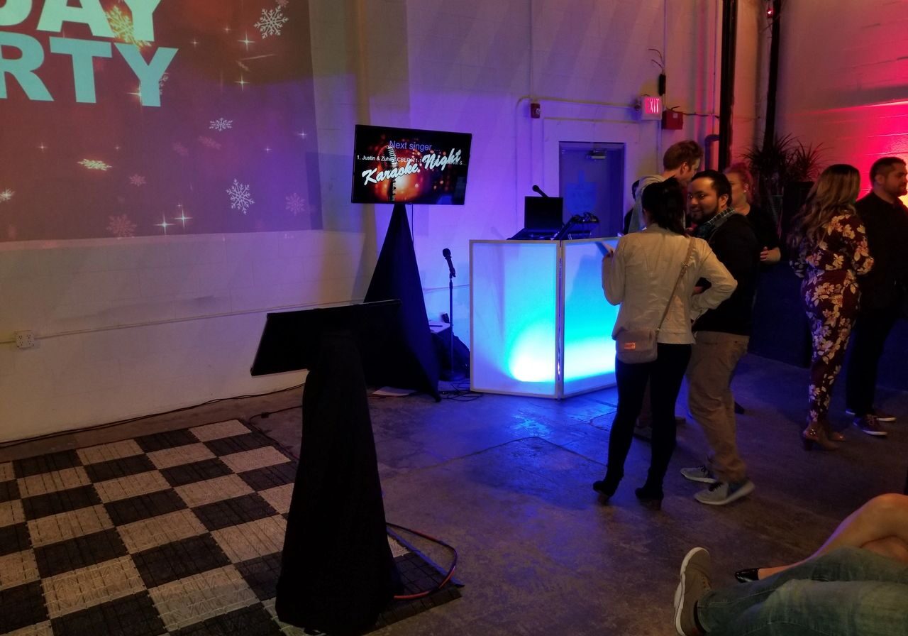 A group of people stands near a brightly lit DJ booth and karaoke setup in an indoor event space with party-themed decorations.