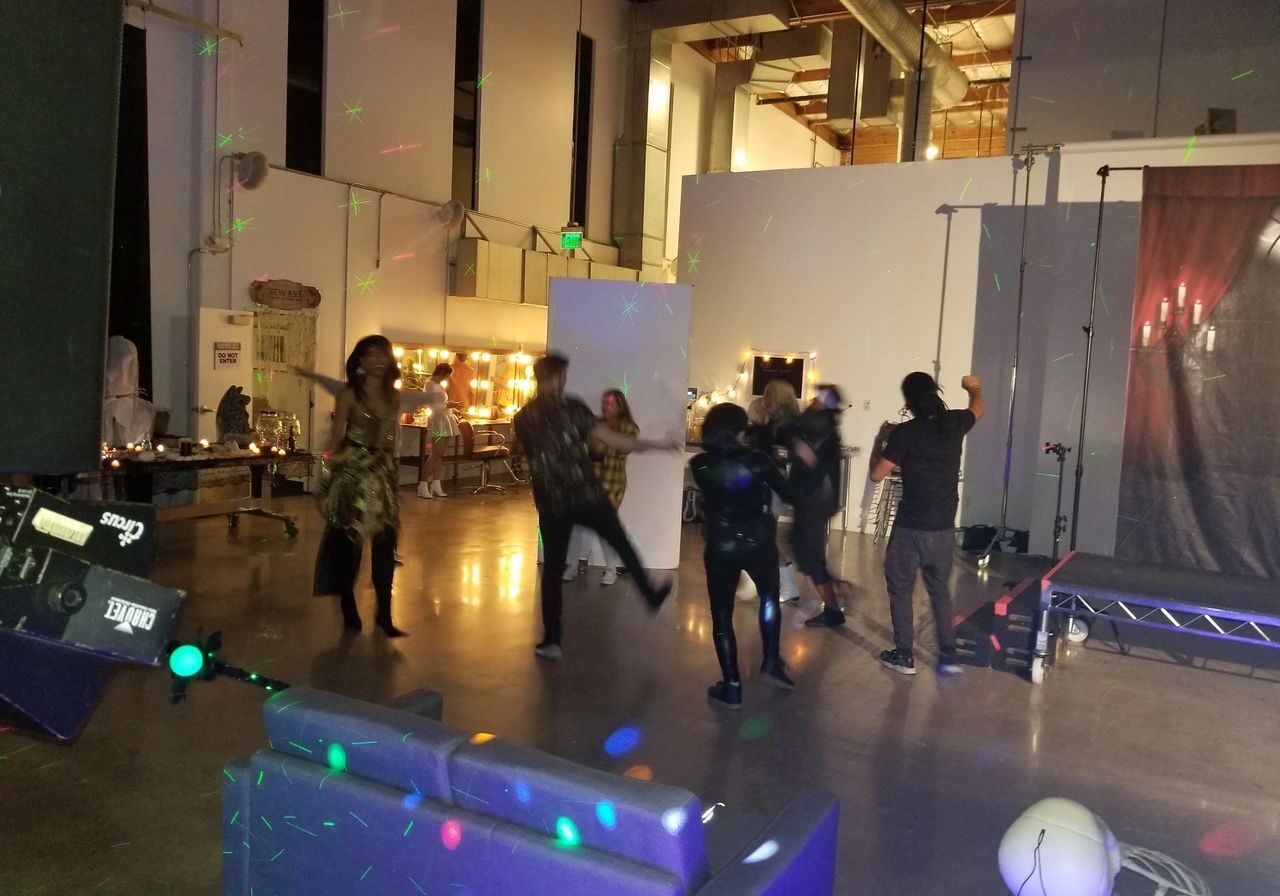 A group of people dance in a dimly lit room with colorful light spots, near a stage and lounge area, with tables and mirrors in the background.