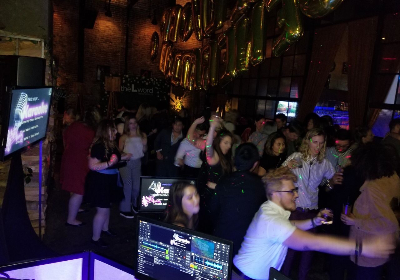 A crowded indoor party with people dancing and socializing under large gold balloons that spell "LOUD"; karaoke lyrics are visible on a screen in the foreground.
