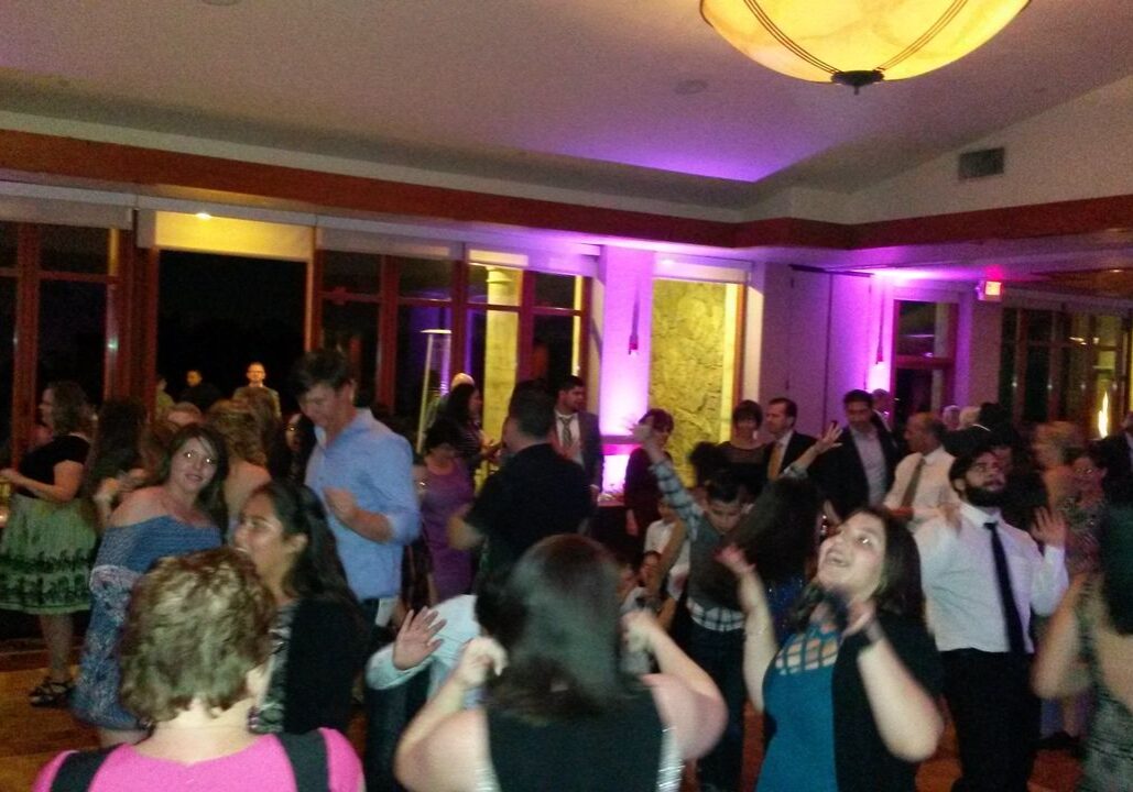 A group of people are dancing and socializing in a brightly lit indoor venue with purple accent lighting.