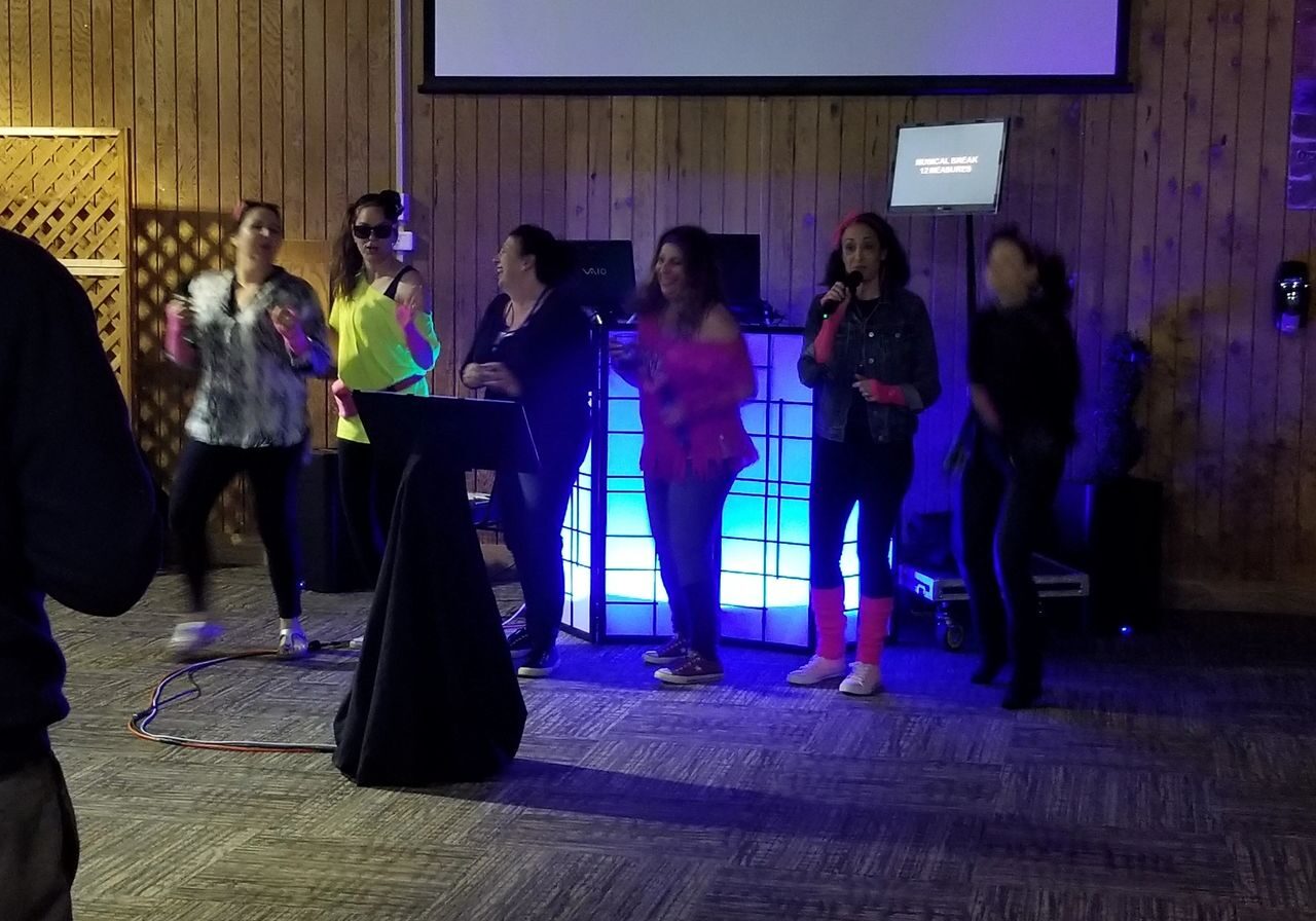 Six people are singing and dancing on a stage in front of a lighted DJ booth and a karaoke screen in a wood-paneled room.