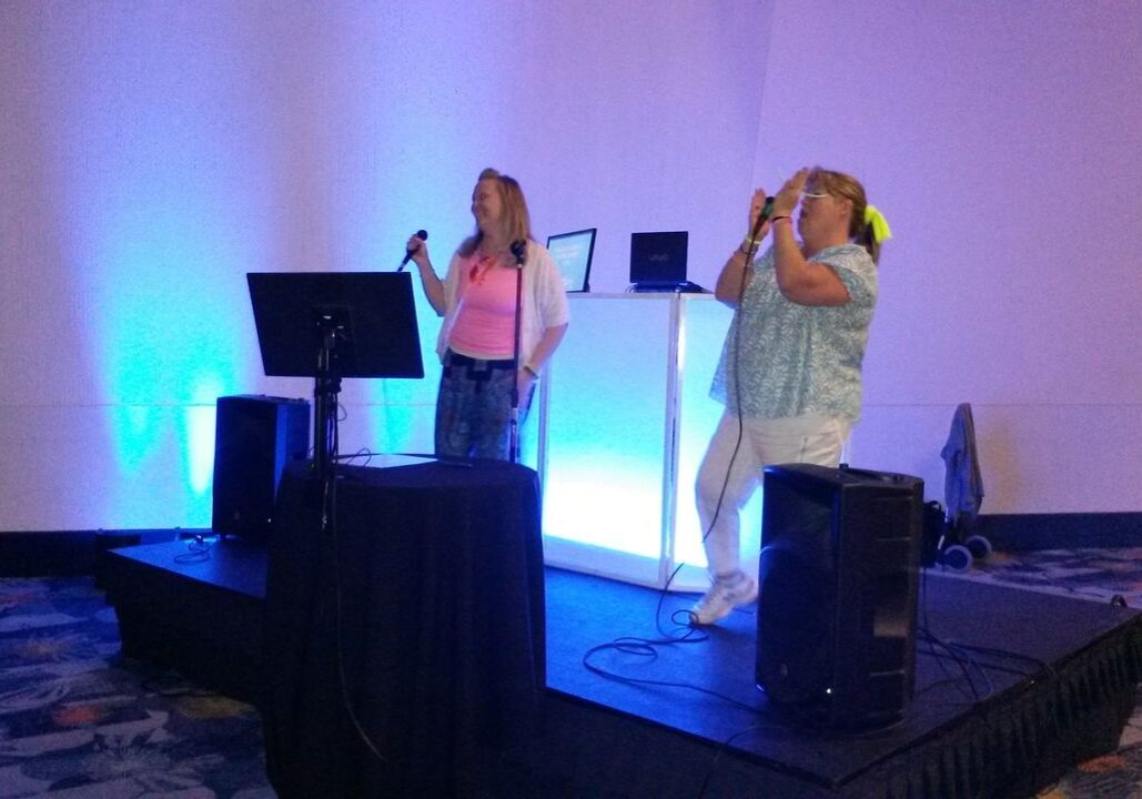 Two women stand on a small stage singing into microphones, with speakers and a screen in front of them and blue lighting behind them.