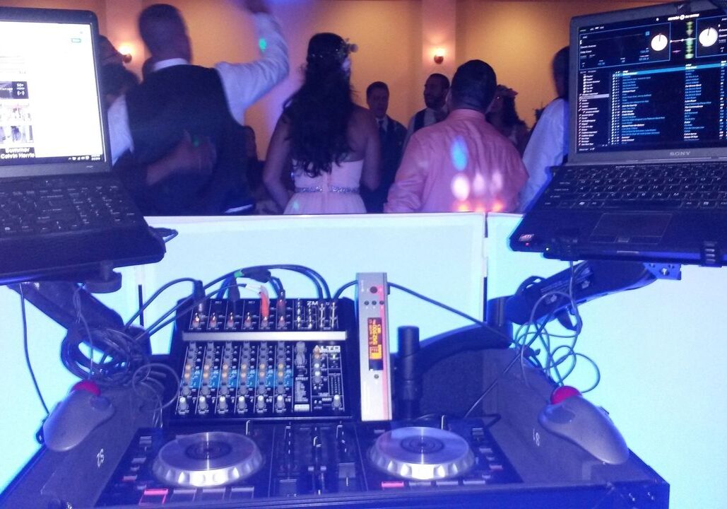 A DJ booth with two laptops and a mixer in the foreground; a group of people dressed formally are dancing in the background.