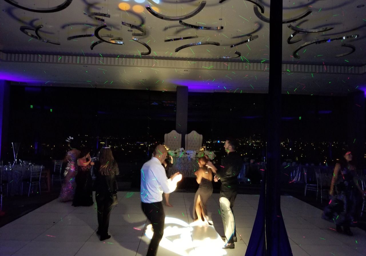 People are dancing on a dimly lit dance floor with colorful lights and cityscape visible through large windows in the background.