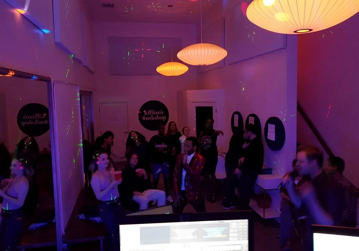 People are gathered in a dimly lit room with colorful lights. Large lamps hang from the ceiling, and a computer setup with monitors is visible in the foreground.