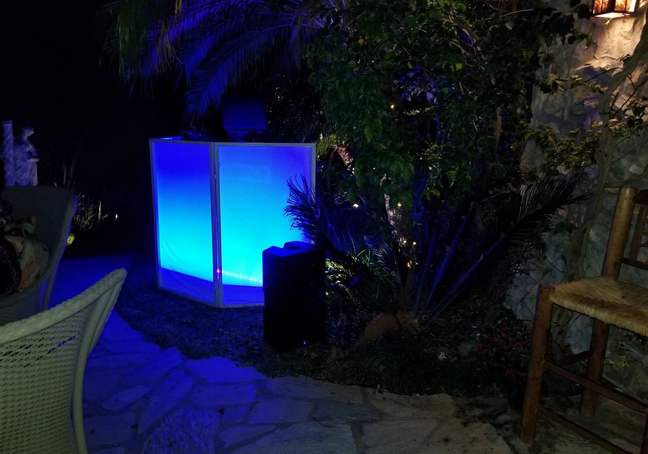 An outdoor scene at night with a glowing blue light panel near tropical plants, a wicker chair, and stone paving visible in the foreground.