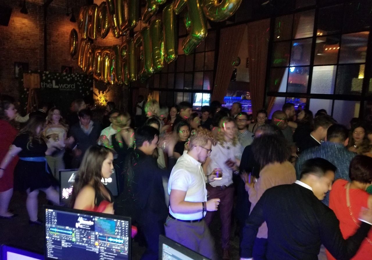 A large group of people are dancing and mingling in a dimly lit indoor venue with a DJ setup in the foreground and gold balloons hanging overhead.