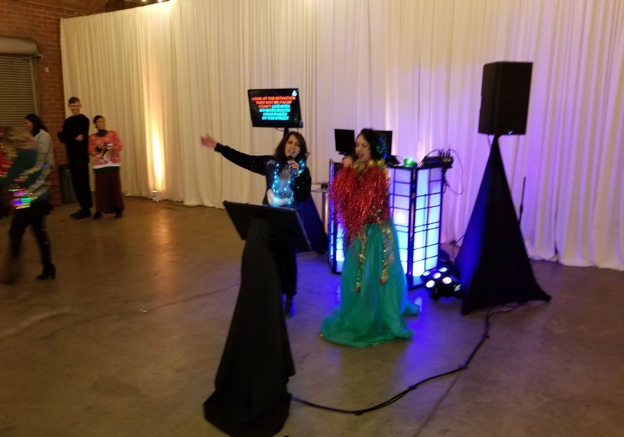 Two people sing karaoke on microphones in front of a screen displaying lyrics, with a white curtain backdrop and DJ equipment nearby at an indoor event.