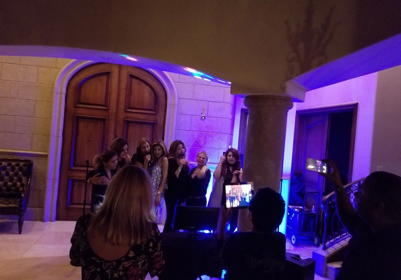 A group of women stand together singing or speaking into microphones in a warmly lit room, while others watch and take photos or videos.
