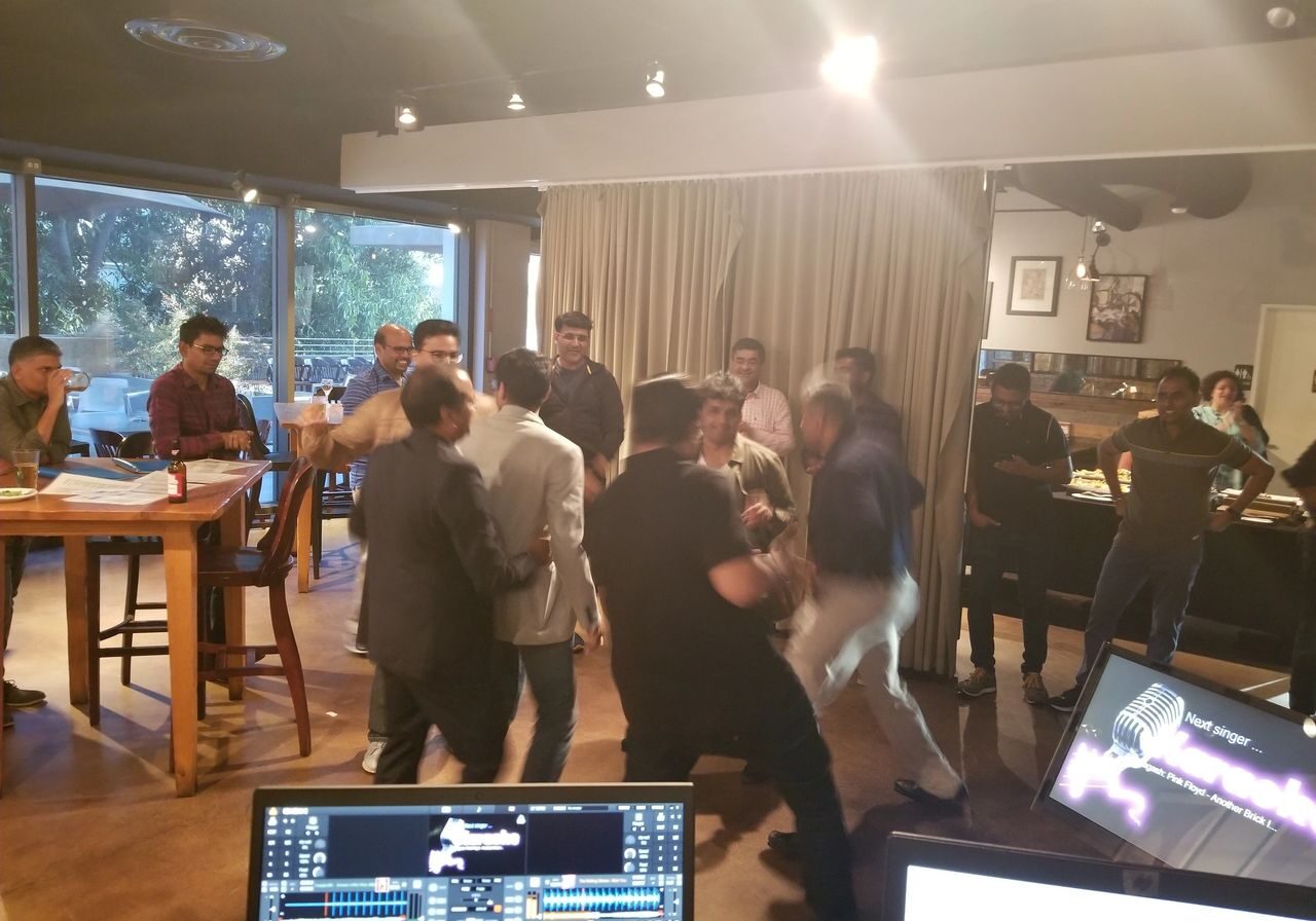 A group of people, some standing and some sitting, gather inside a restaurant; several appear to be involved in an altercation while others watch. Computer monitors are visible in the foreground.