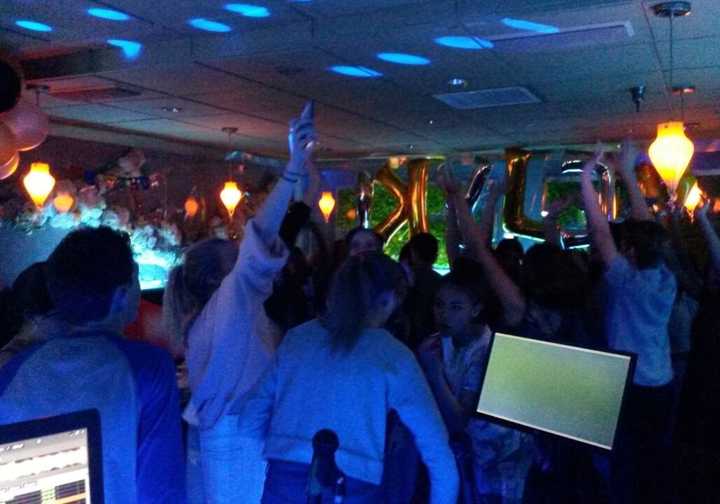 A group of people dance and raise their arms at a dimly lit party with balloons, decorations, and blue lighting.