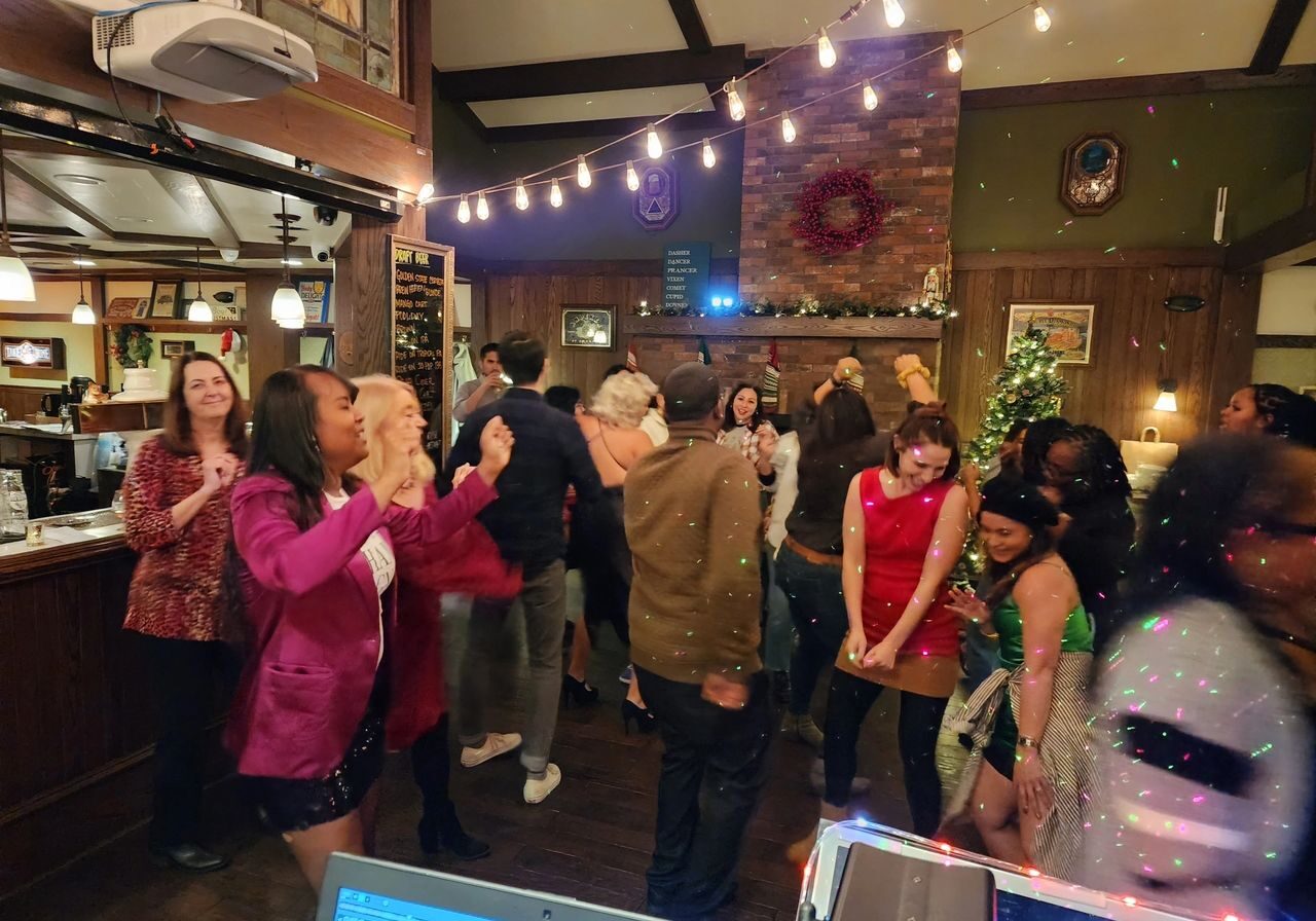 A group of people dance and socialize in a warmly lit indoor venue decorated with string lights and holiday ornaments.