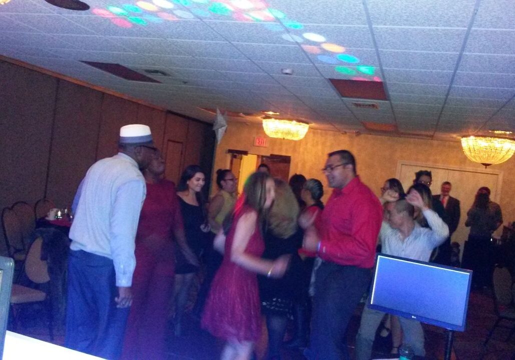 A group of people are dancing in a dimly lit event room with chandeliers, while others sit at tables. A DJ booth and computer screens are visible in the foreground.