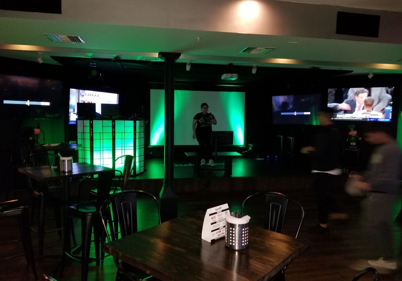 Dimly lit bar with empty tables and chairs, green-lit stage in the center, one person on stage, several TV screens on surrounding walls, and a few people moving around.