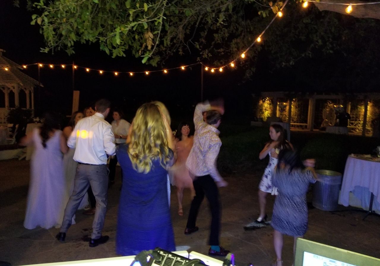A group of people dance outdoors at night under string lights, with trees and a table visible in the background.