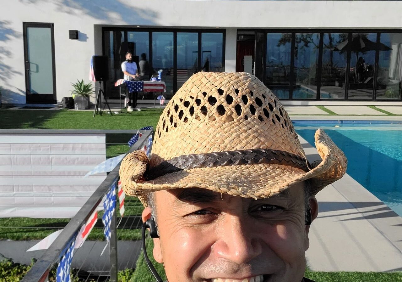 A man in a straw hat smiles for a selfie by a pool, with a modern house, American flags, and a party DJ playing music near a table in the background.