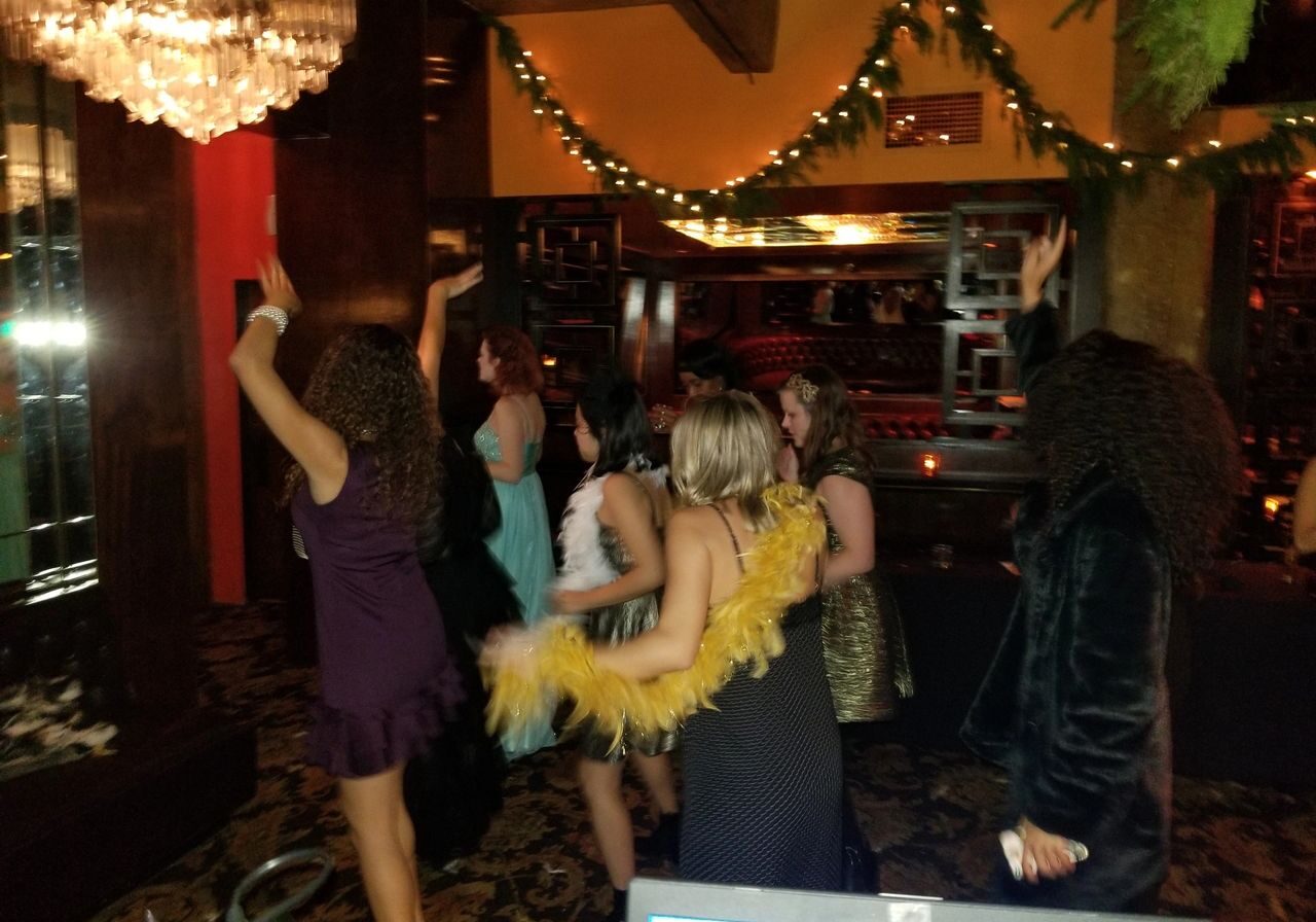 A group of women in dresses and costumes dance and raise their arms in a dimly lit, festively decorated room.