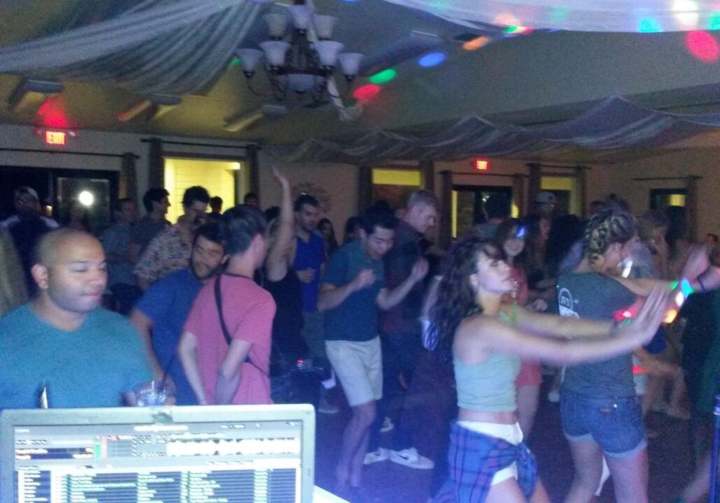 A crowd of people dance in a dimly lit indoor venue with colorful lights, as a DJ booth is visible in the foreground.
